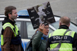 Ein Plakat zeigt ein weißes Lamm, das mit »our children« beschriftet ist. An dem Lamm reißen schwarze Wölfe, unter anderem mit der Aufschrift »NWO«, »MSM«, »pedophilia« und »LGTBQ Propaganda« . Die Aktivistin, die es trägt, unterhält sich mit einem Polizisten
